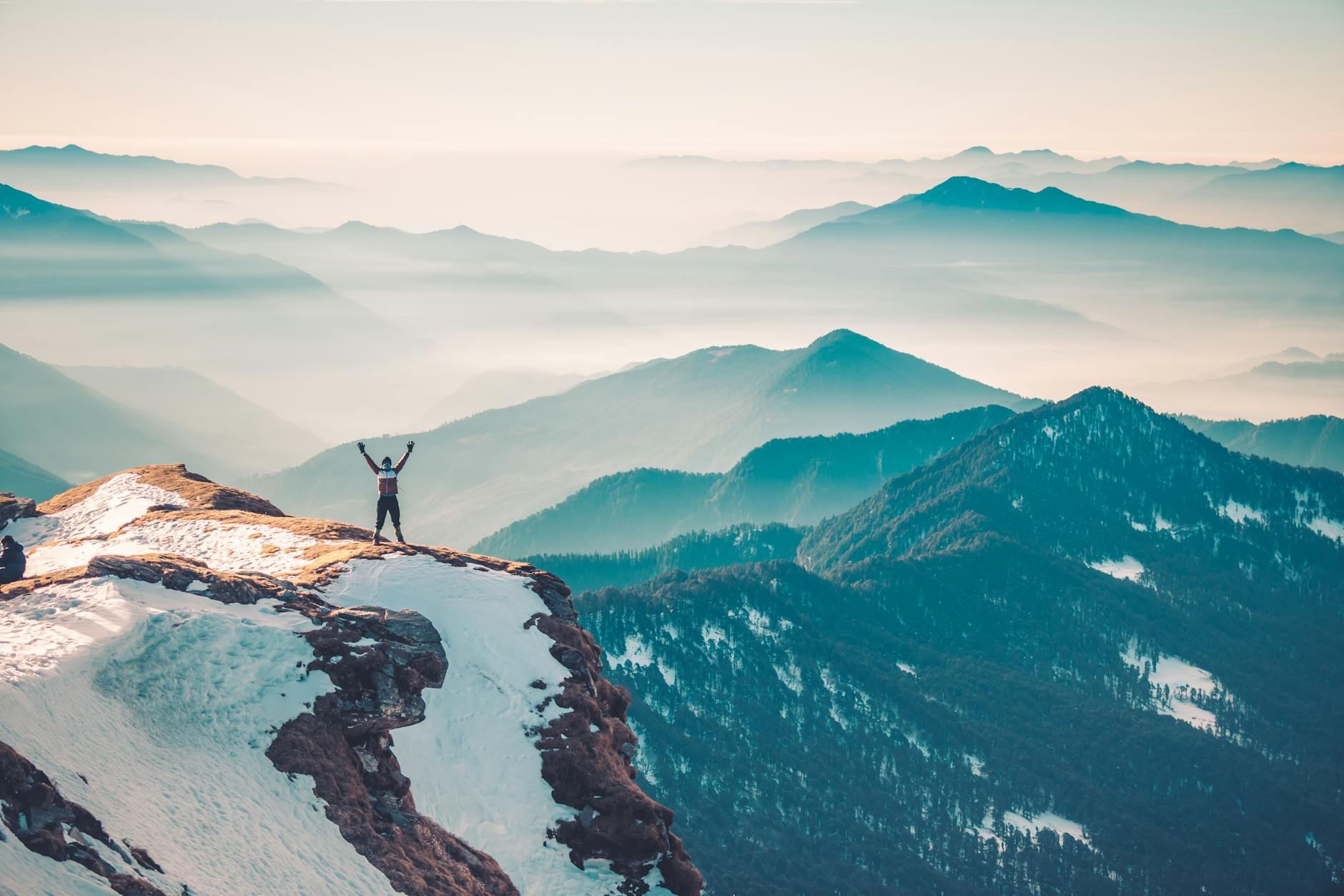 A hiker stands on a snowy mountain peak, arms raised in triumph, surrounded by a vast mountain range.