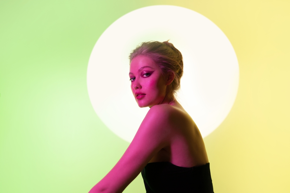 Fashion art portrait of woman in neon color light looking to the camera on green neon background. Behind the girl on the wall is a circle from a spotlight.
