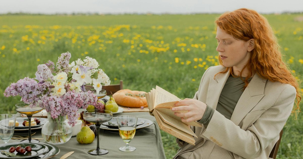 Woman with red hair reading at a beautifully set outdoor picnic in a summer field.