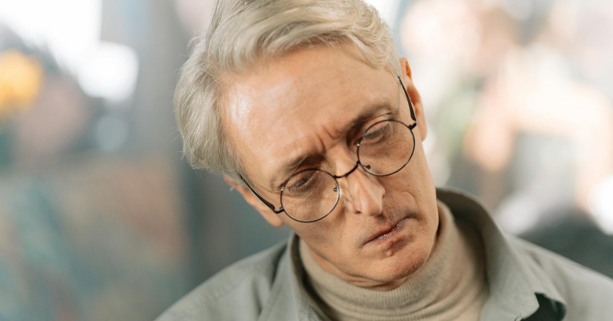 Elderly man with glasses and gray hair appearing pensive and thoughtful indoors.