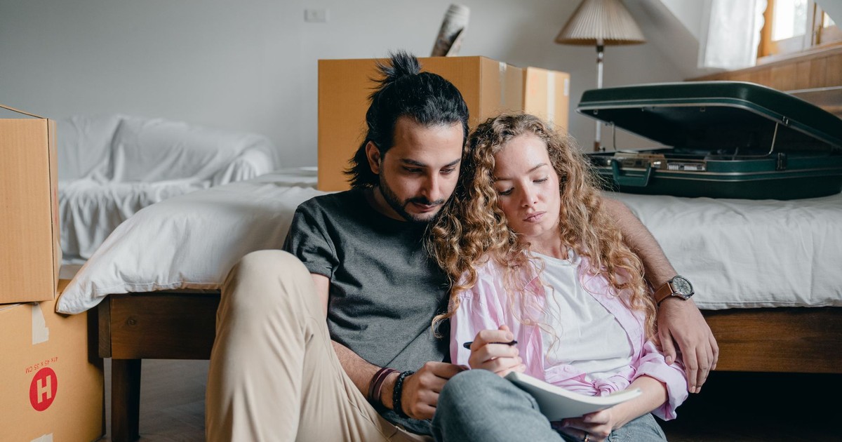 Thoughtful male and female in casual wear sitting near bed among boxes together and taking notes while moving house