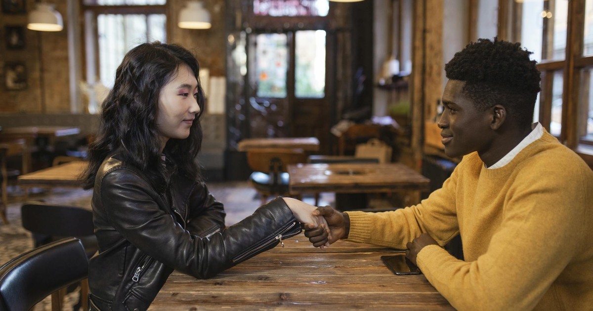 A diverse man and woman sit in a café, shaking hands and smiling warmly.