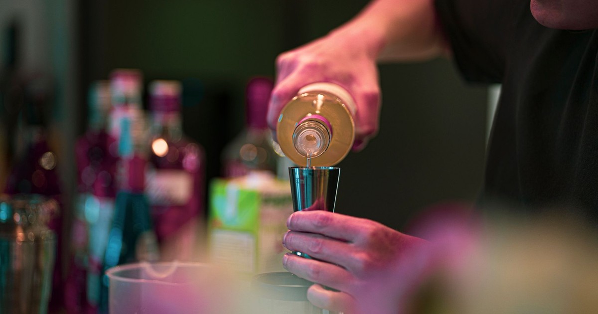 A bartender pours a drink into a jigger at a stylish bar with bottles in the background.