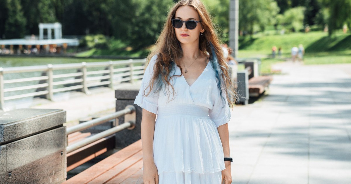 A young woman in a white dress and sunglasses enjoys a sunny day outdoors.