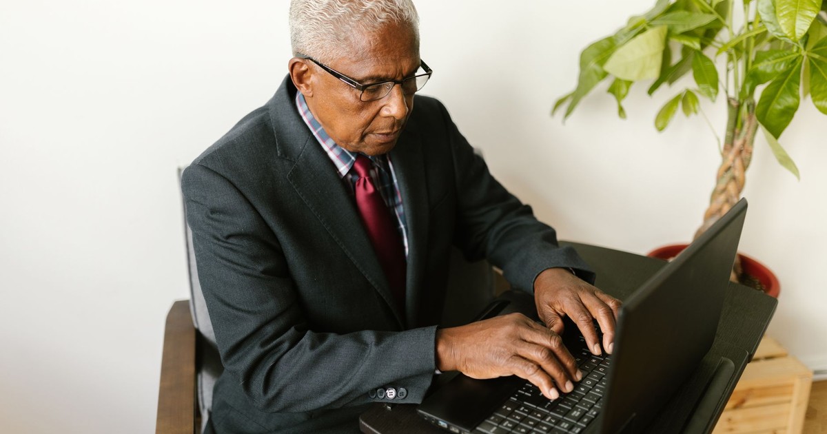 Elderly man in a gray suit working on a laptop at an office desk.