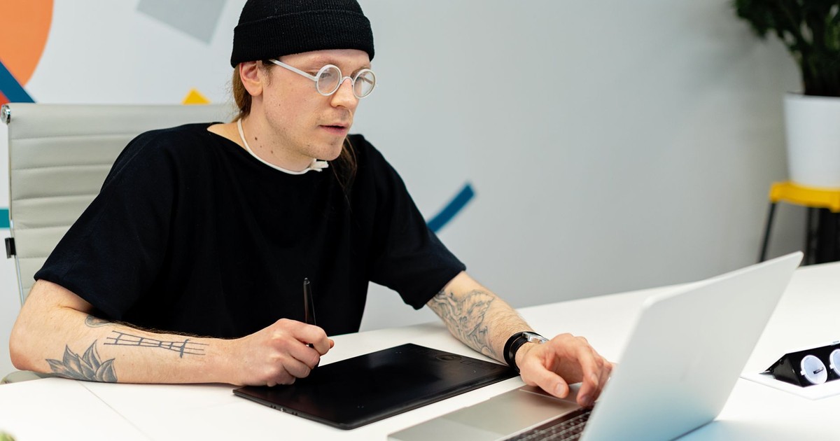 Tattooed designer working on a laptop and tablet in a stylish office setting.