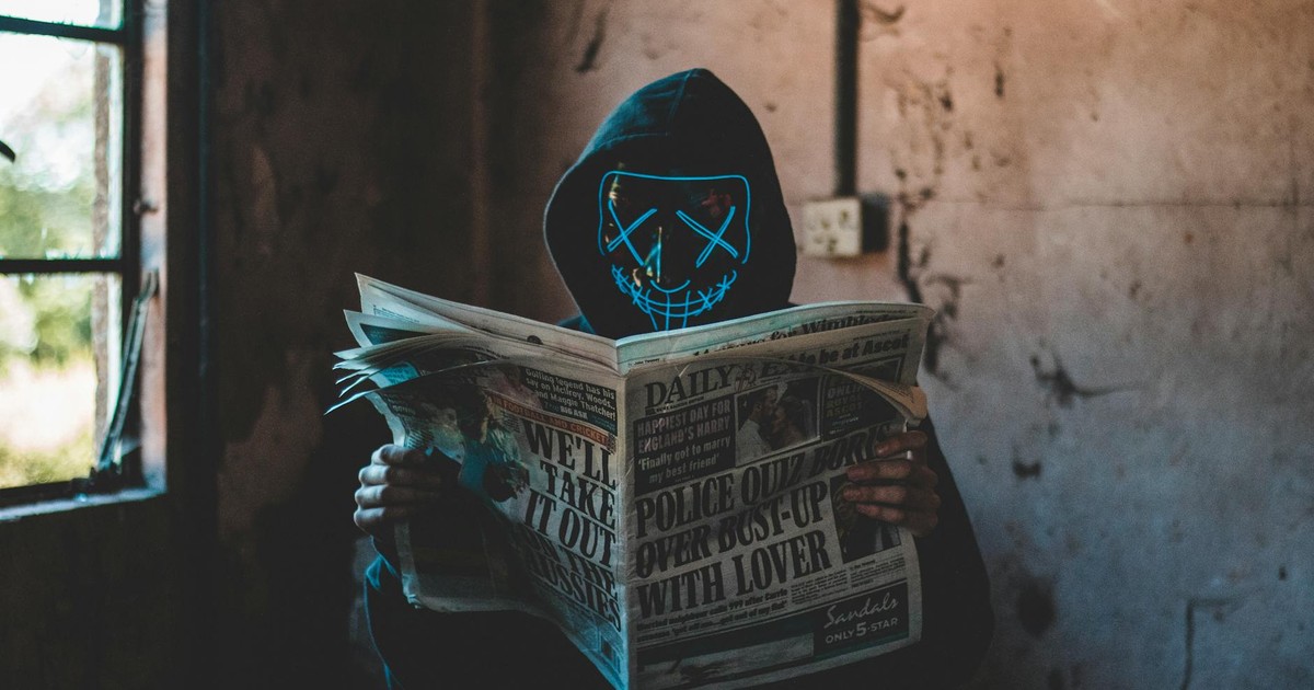 A person in a neon mask and hoodie reads a newspaper in an eerie, abandoned room.