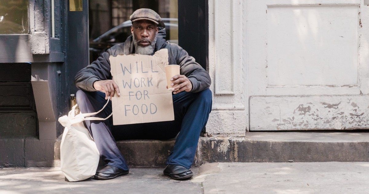 A man sitting on a sidewalk holding a 'Will Work for Food' sign, depicting urban hardship.