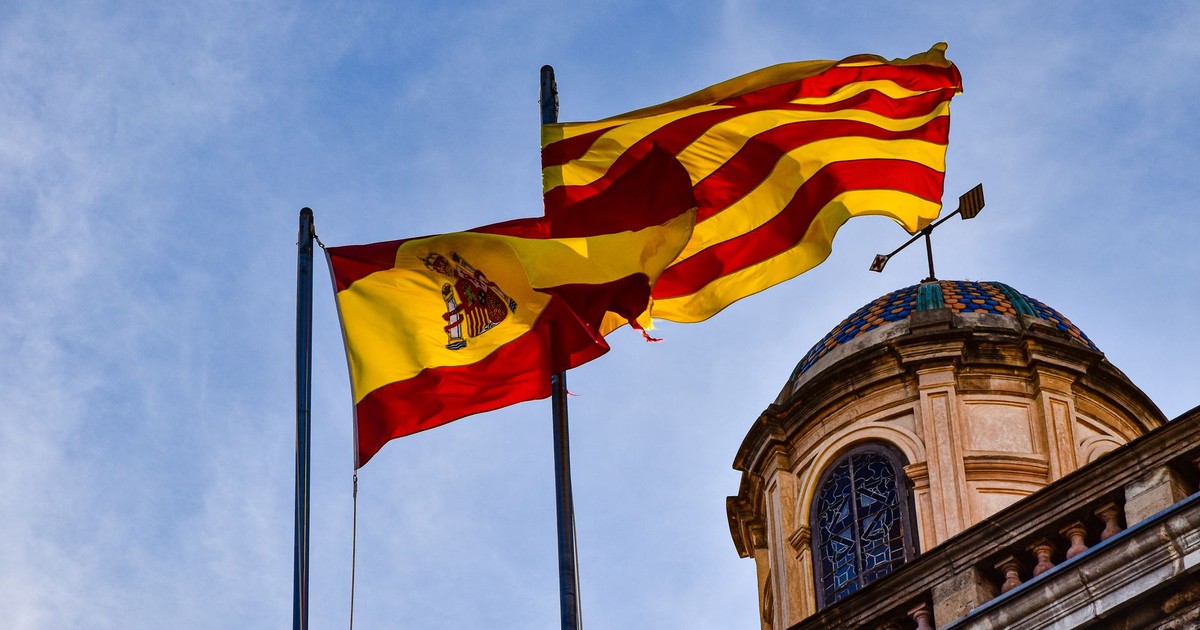 Spanish and Catalan flags waving against a blue sky in front of a historical building.