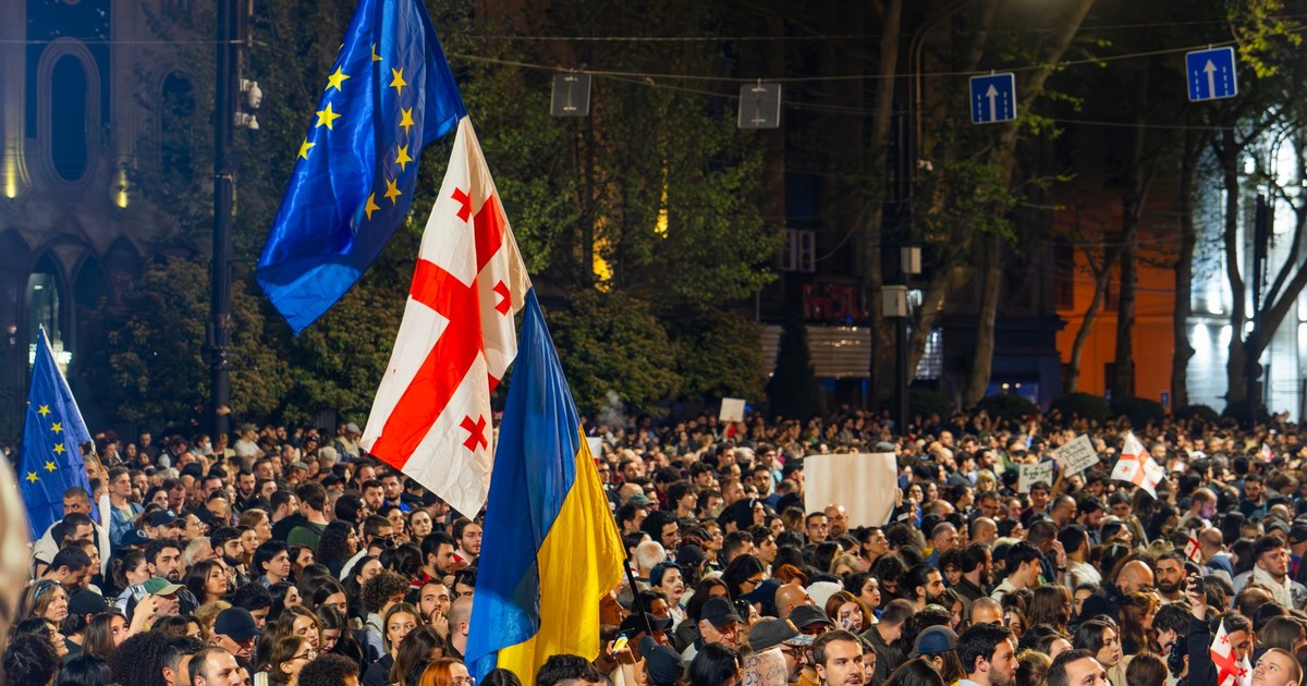 Crowd of protesters in Tbilisi holding Georgian, EU, and Ukrainian flags at night.