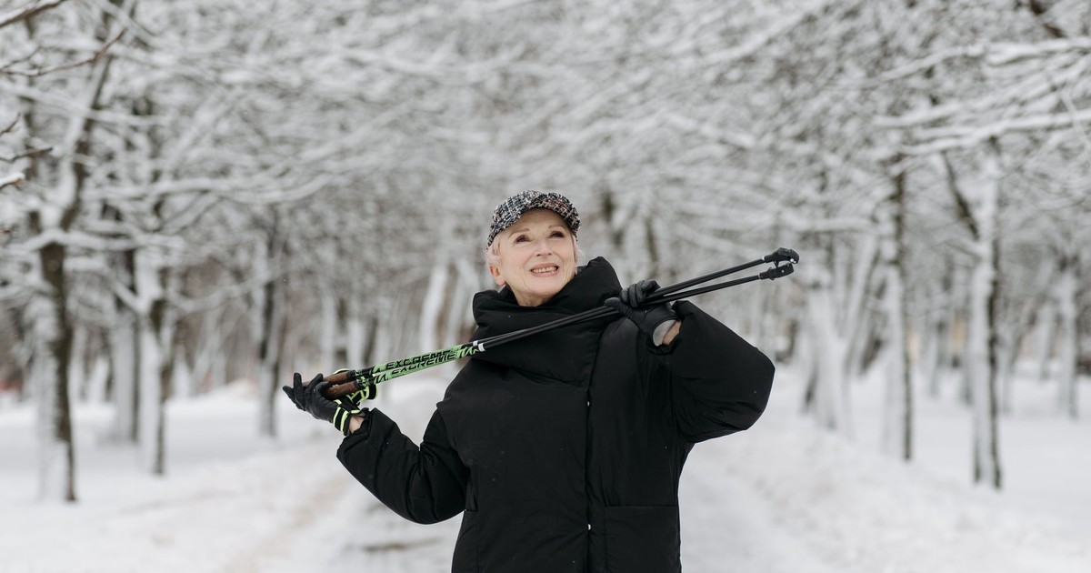 A senior woman smiling while hiking with trekking poles in a snowy forest.