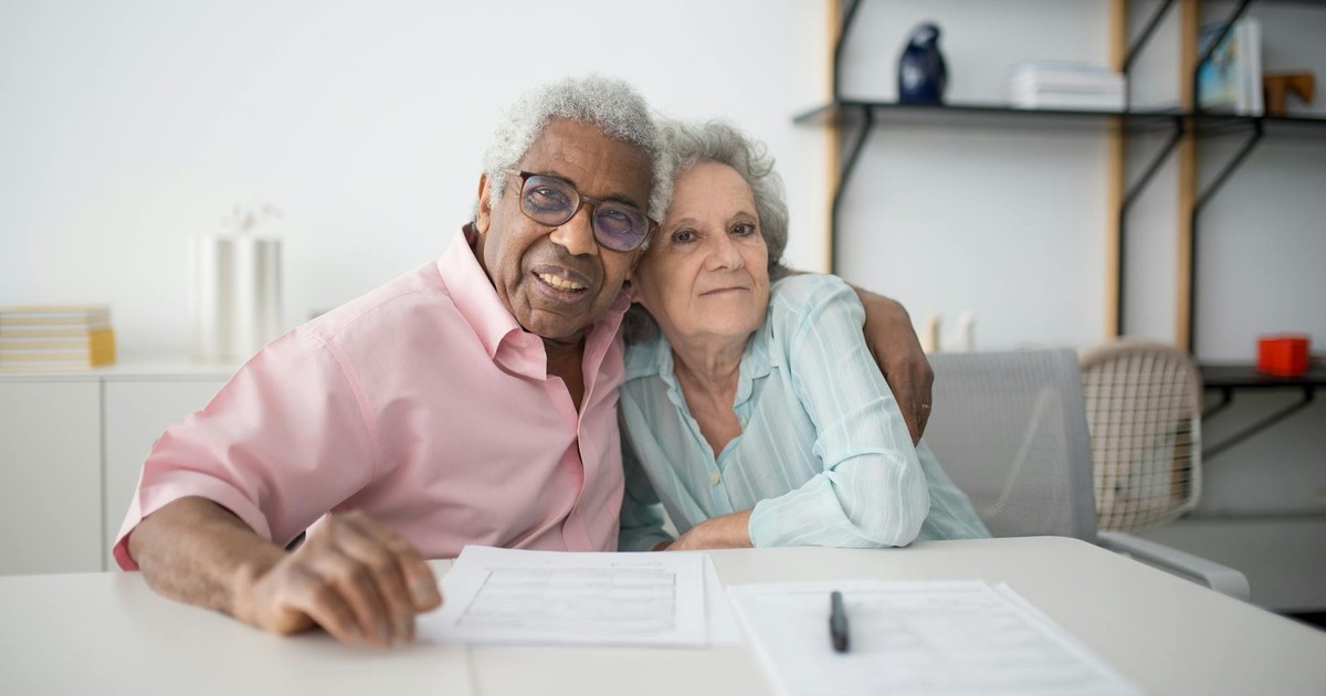 Elderly couple smiling and hugging while looking at documents on table indoors.