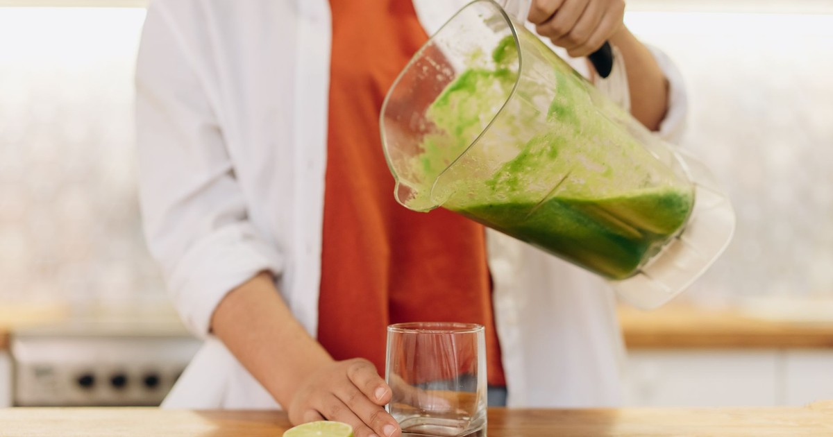 Close-up of a person pouring a vibrant green smoothie into a glass with limes on the wooden table.