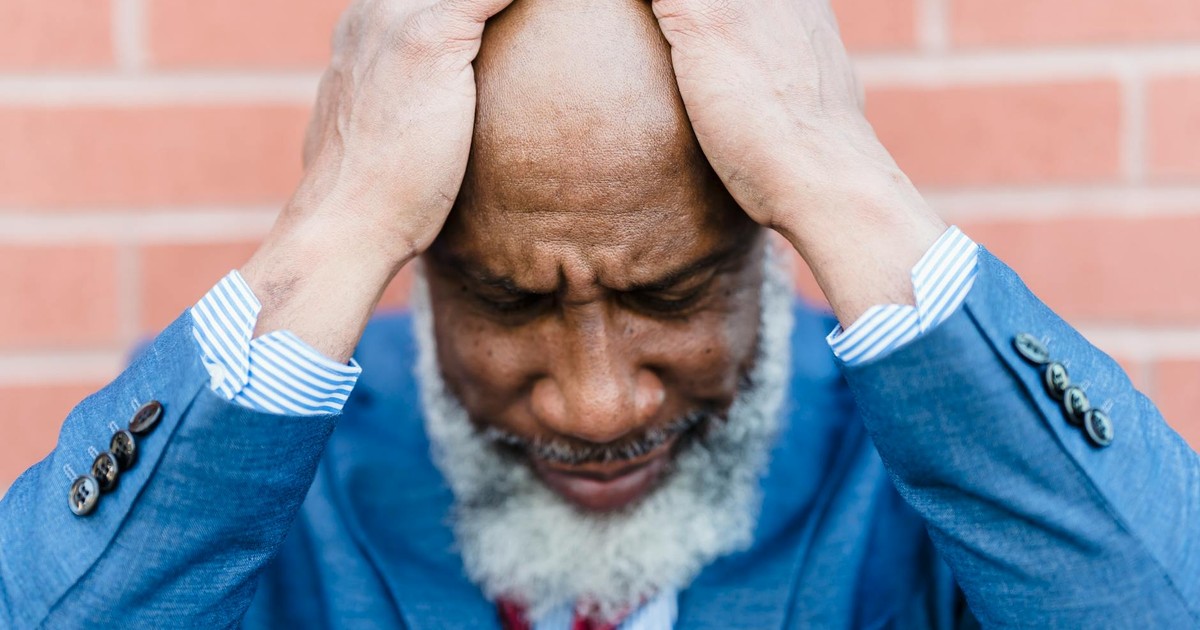 Unhappy bald African American male worker with gray beard in classy wear standing near building with hands on head on street