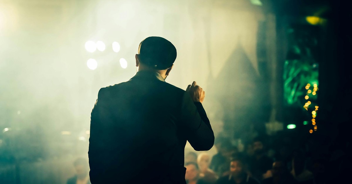A man seen from the back speaking to an audience at an atmospheric entertainment venue.