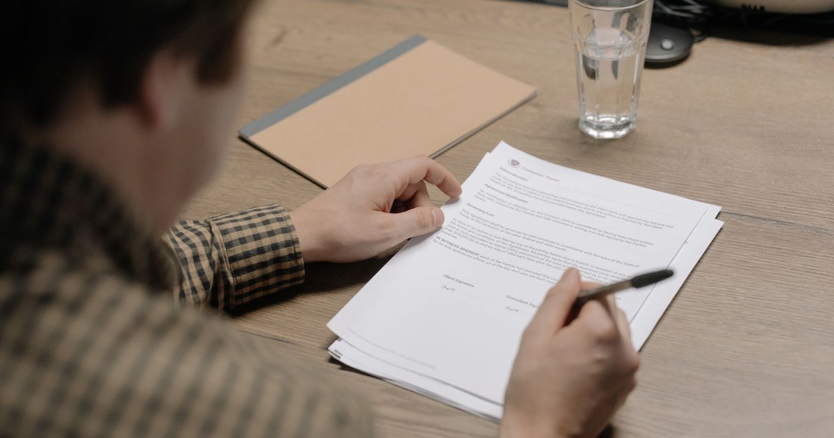 A businessman is reviewing a contract while holding a pen at a desk with a glass of water nearby.