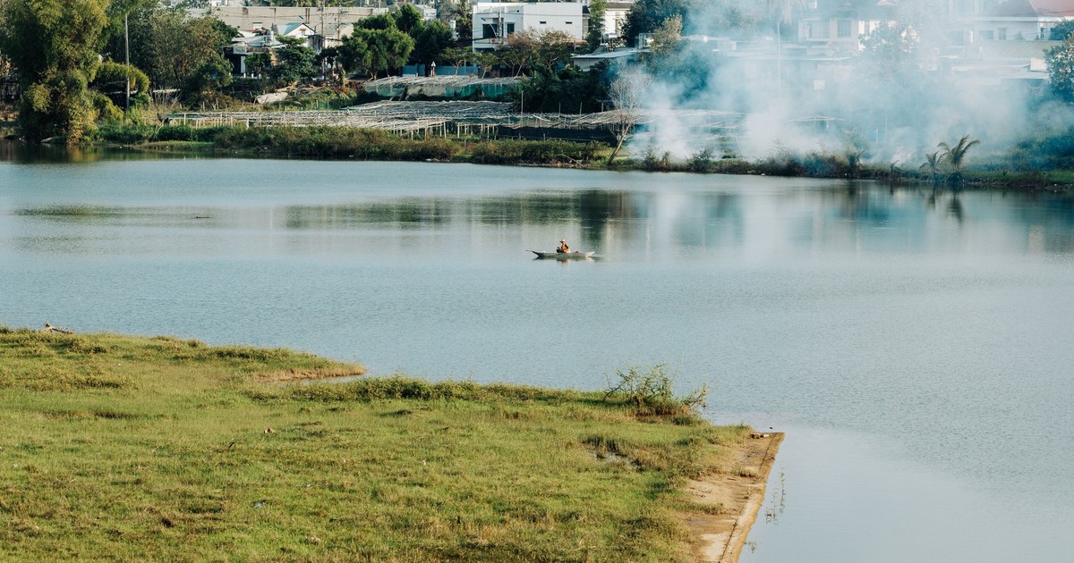 Peaceful lake view with lone canoeist in Kon Tum, Vietnam, framed by greenery and smoke in the distance.