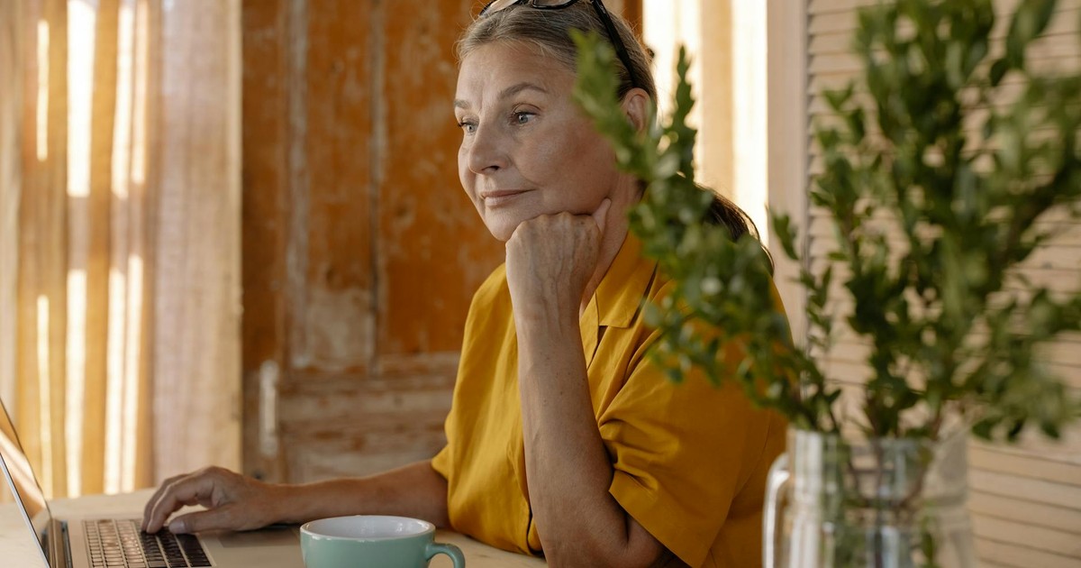 Elderly woman sitting at a table using a laptop, relaxing in a cozy home setting.
