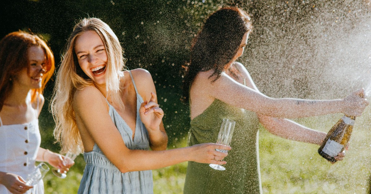 Three women enjoying a sunny day, laughing and celebrating with champagne outdoors.