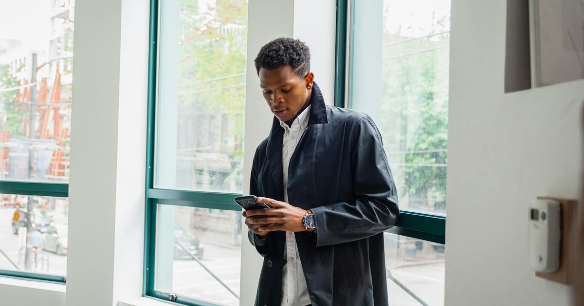Businessman in a black jacket using smartphone indoors by office window. Modern and professional setting.