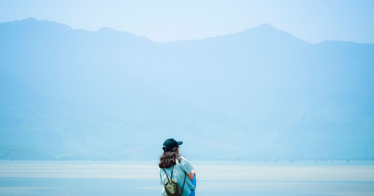 A person sitting alone by a lake, with mountains in the background, under a bright sky.