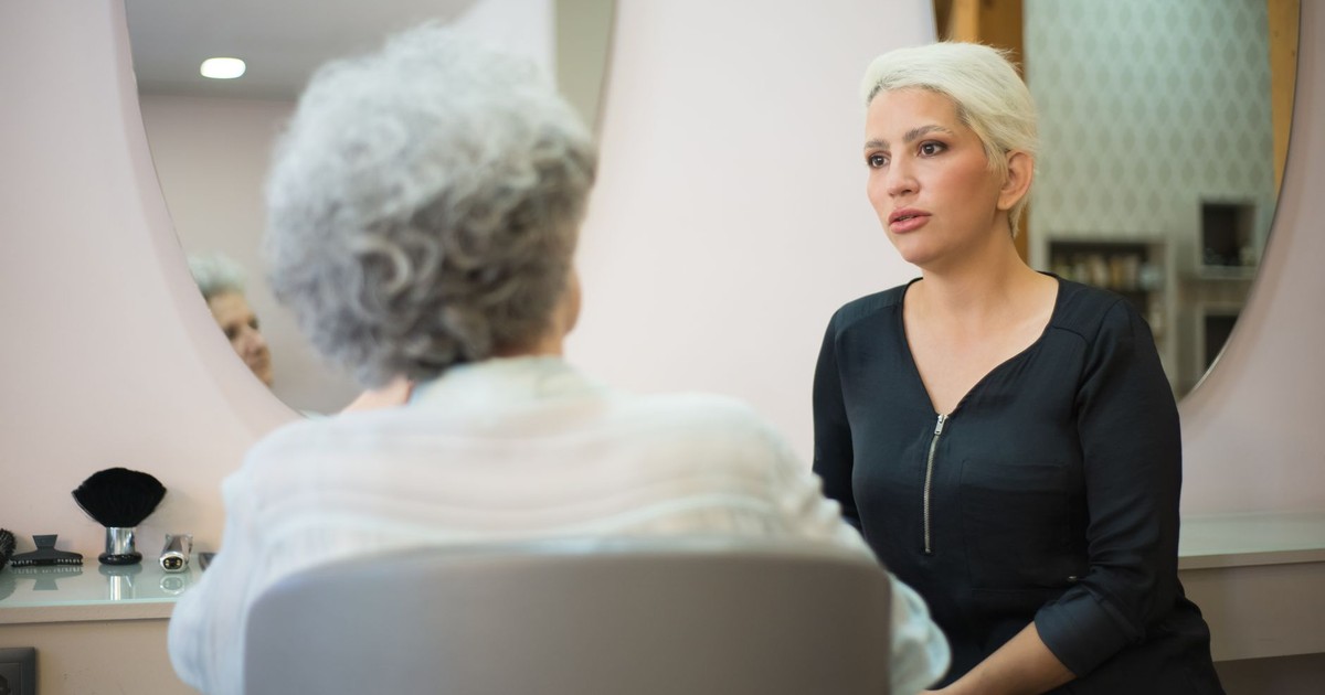Two women having a conversation in a salon setting with a mirror reflecting their interaction.
