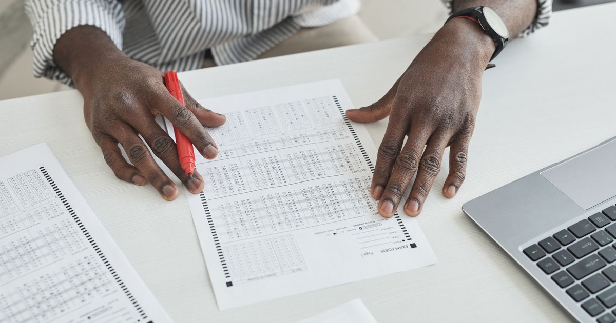 An educator carefully checks exam papers using a red marker at a desk, illustrating focused grading.