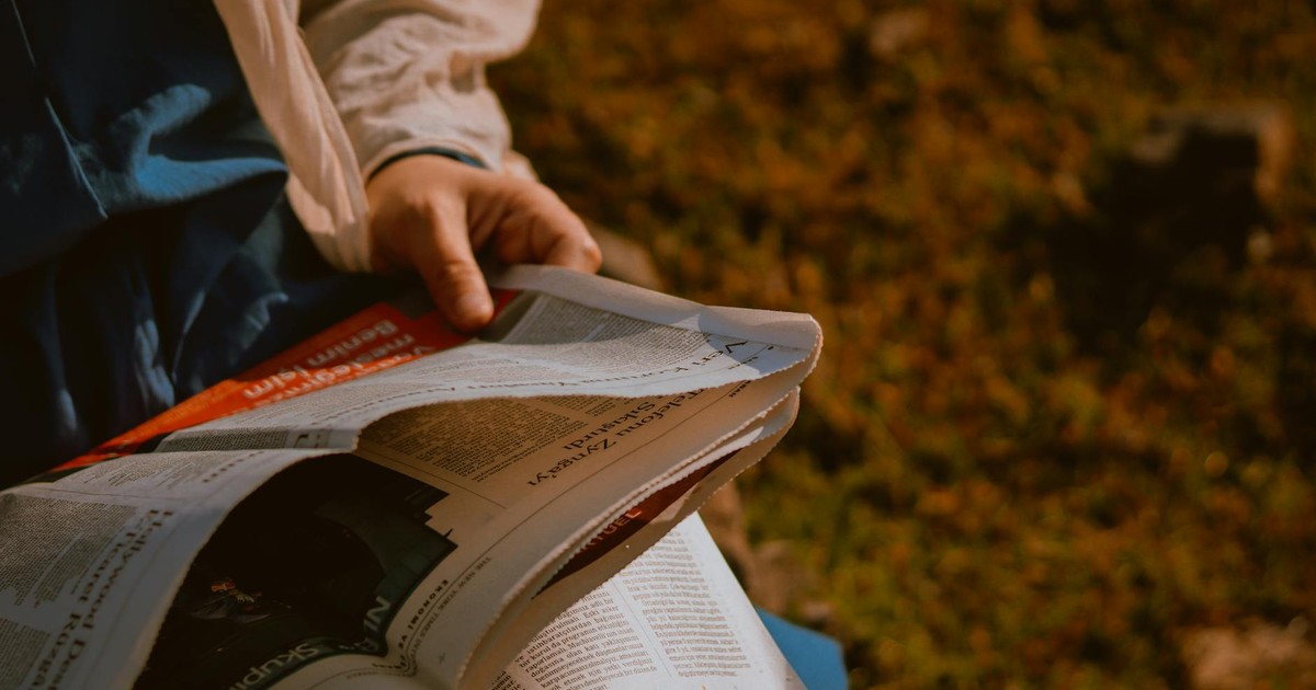 From above of crop unrecognizable person reading newspaper on green meadow in daylight