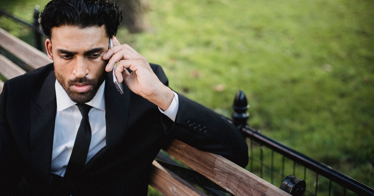 A man in a black suit sitting on a bench, engaged in a serious phone conversation in a park.