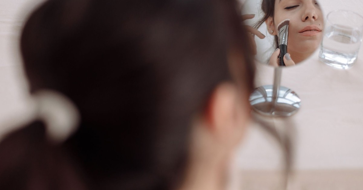 A woman applying makeup using a brush while looking into a small mirror.