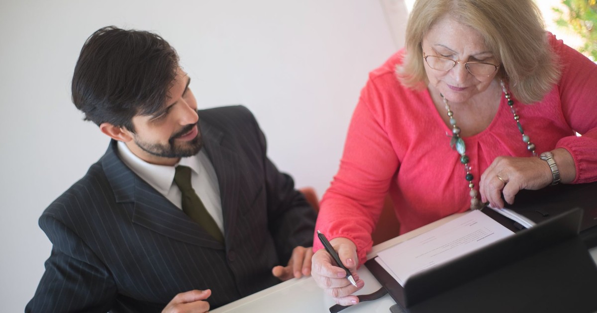 A senior woman signing paperwork with a young professional assisting her at a desk.