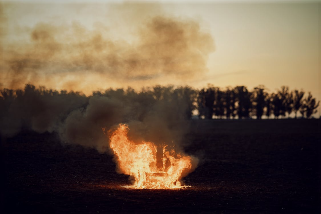 A solitary bonfire burns intensely on a vast open field, silhouetted by the evening sky