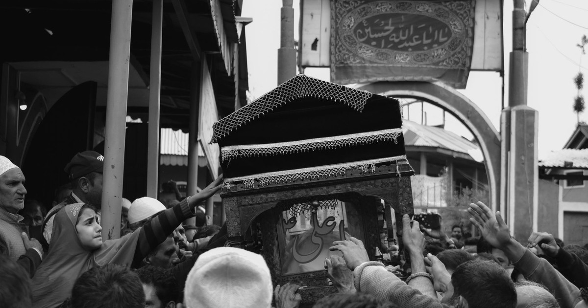 A monochrome image of a crowd carrying a decorated casket during an outdoor funeral procession.