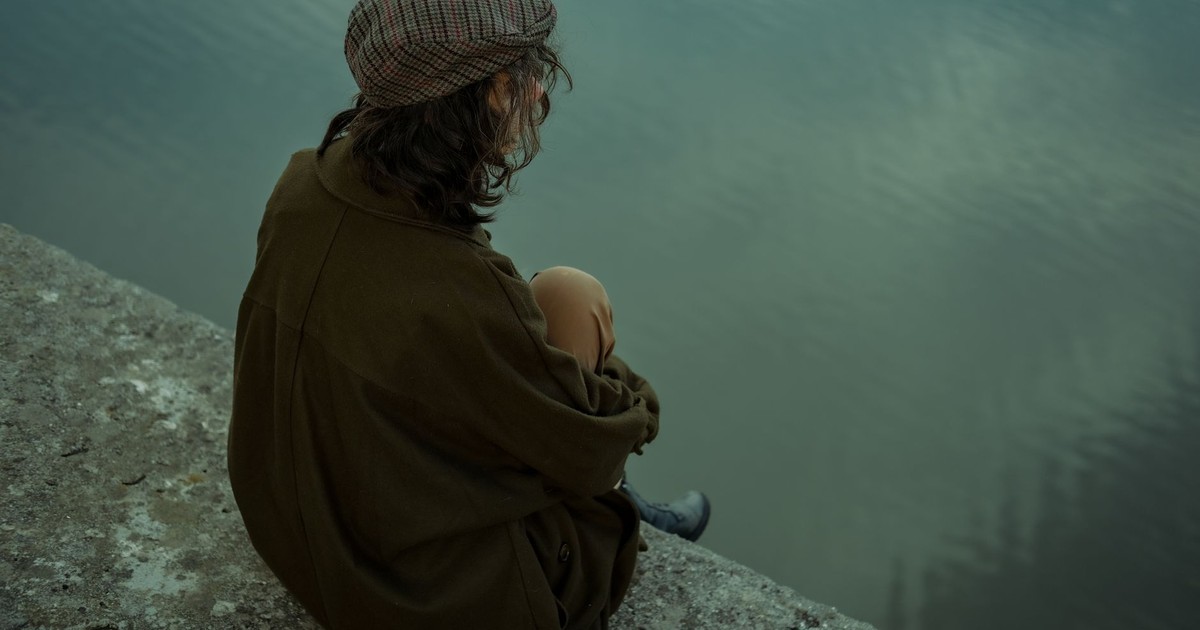 A woman in casual attire sits thoughtfully by the lake, evoking calm and introspection.