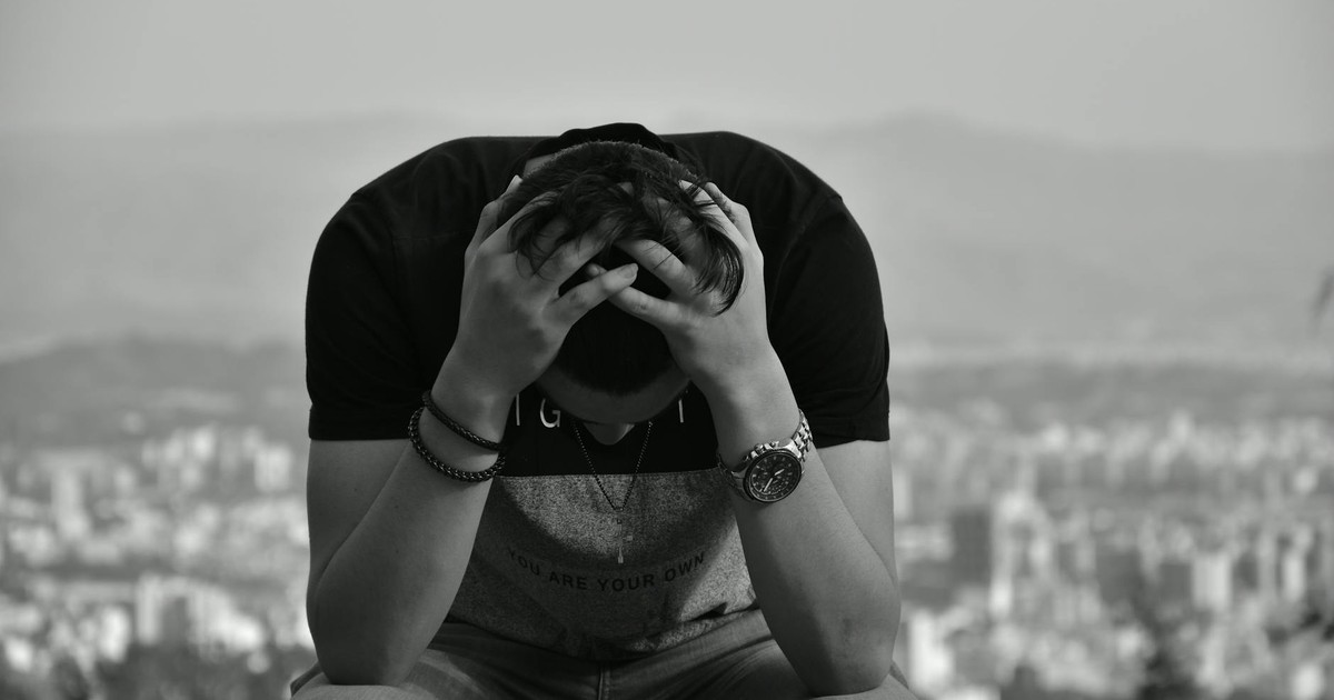 A focused young man sits with hands on head against an urban cityscape in grayscale, suggesting introspection.