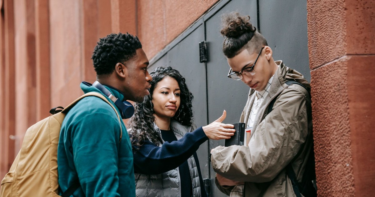 Multiracial young students in casual clothes with backpacks standing in city street while bullying male with books