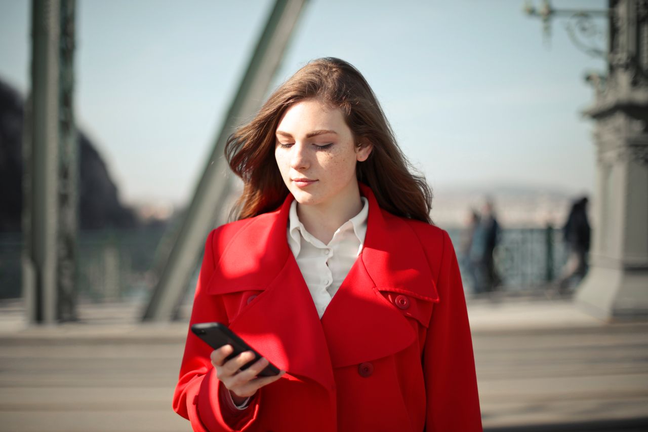 Young woman in a vibrant red coat looking at her smartphone while standing outdoors on a city bridge
