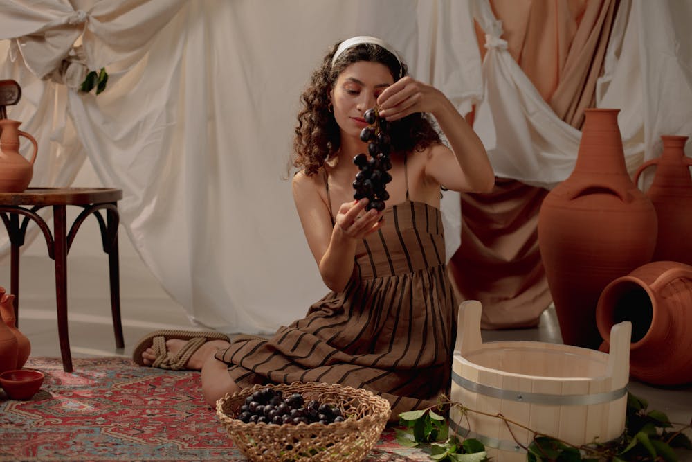 Woman holding grapes in a Roman-inspired indoor setting, surrounded by pottery and draped fabric.