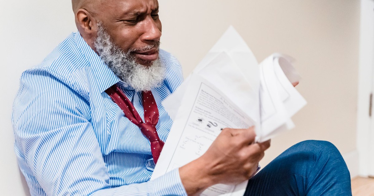 African American man in distress with paperwork, sitting against a wall indoors.