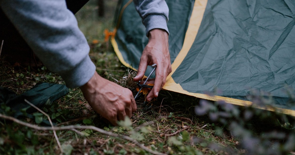 A close-up of a person securing a tent peg in a forested campsite, depicting outdoor camping activities.