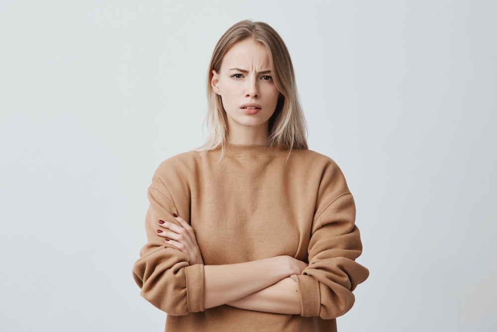 Waist-up portrait of beautiful girl with blonde straight hair frowning her face in displeasure, wearing loose long-sleeved sweater, keeping arms folded. Attractive young woman in closed posture.