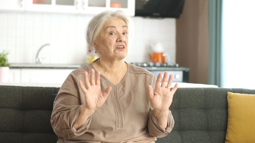Unhappy and grumpy old woman is resting in her quiet home. White-haired old woman sitting alone on the sofa talking to the camera.