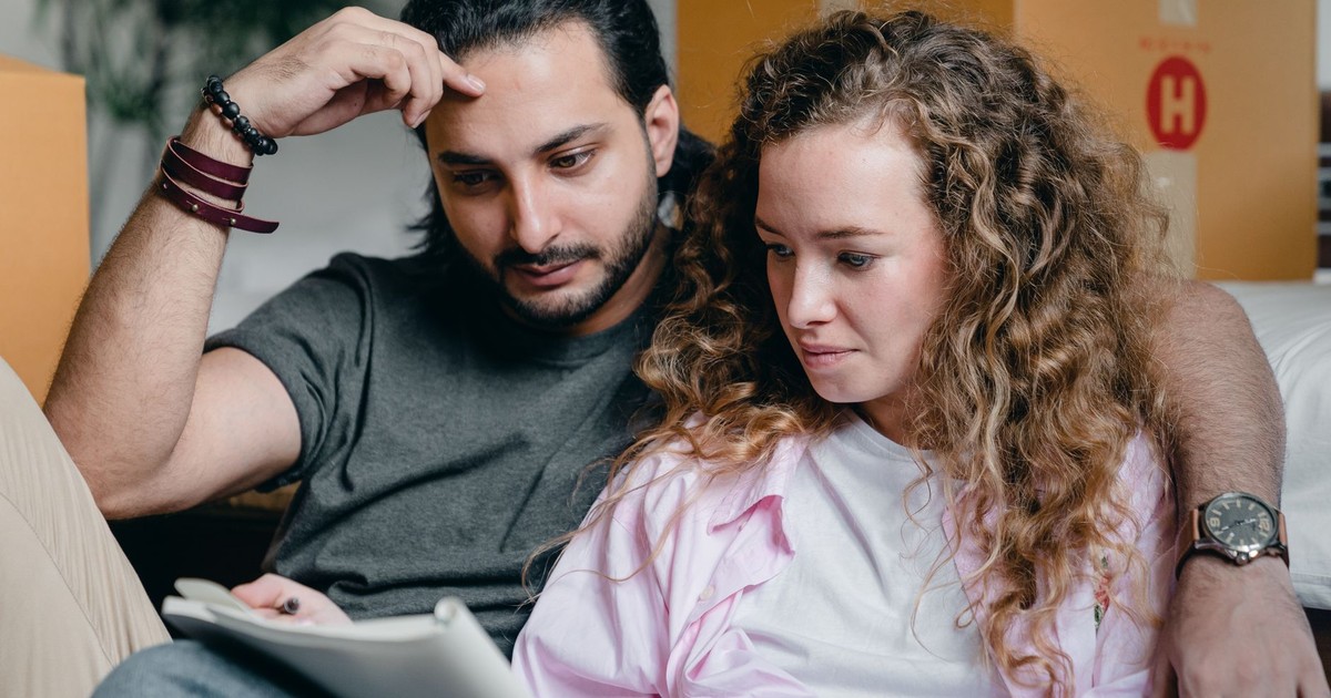 Focused male and female in casual wear sitting near boxes and looking through notebook thoughtfully while moving house