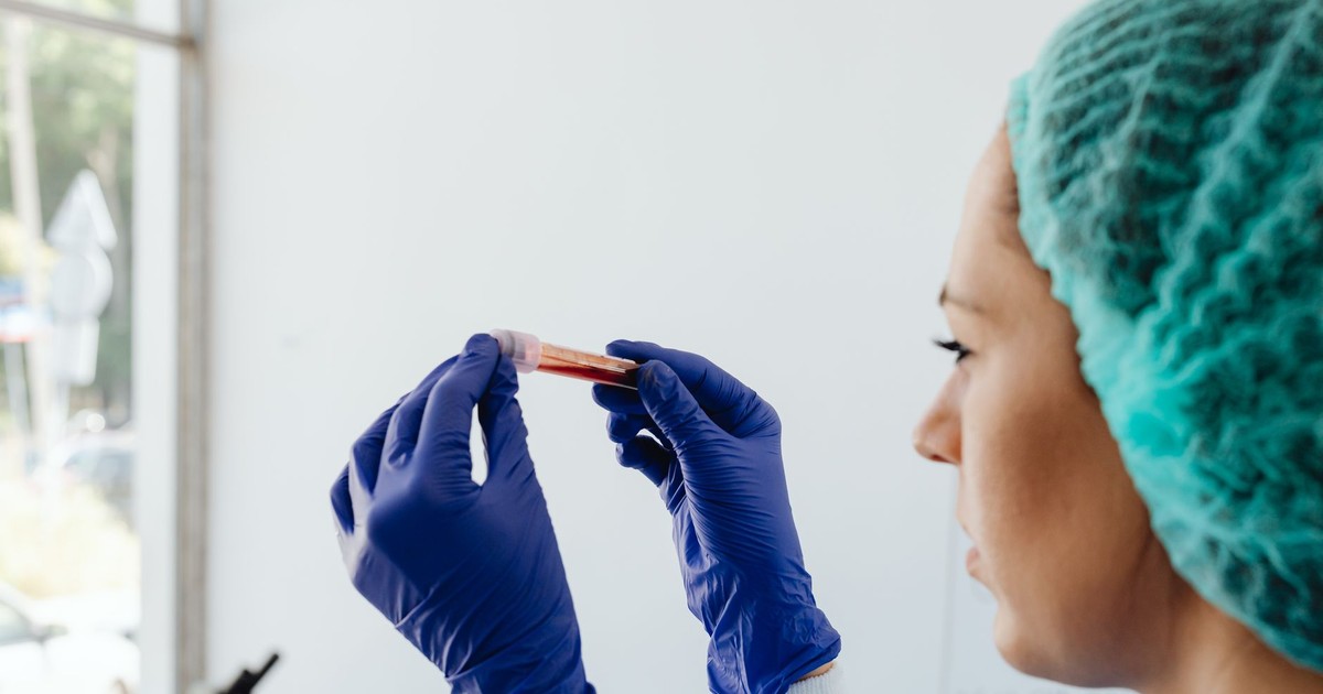 A focused lab technician holds a blood sample in a test tube, wearing gloves and a hair net.