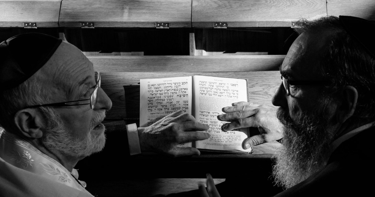 Two elderly men discussing Hebrew text in a synagogue, captured in black and white.