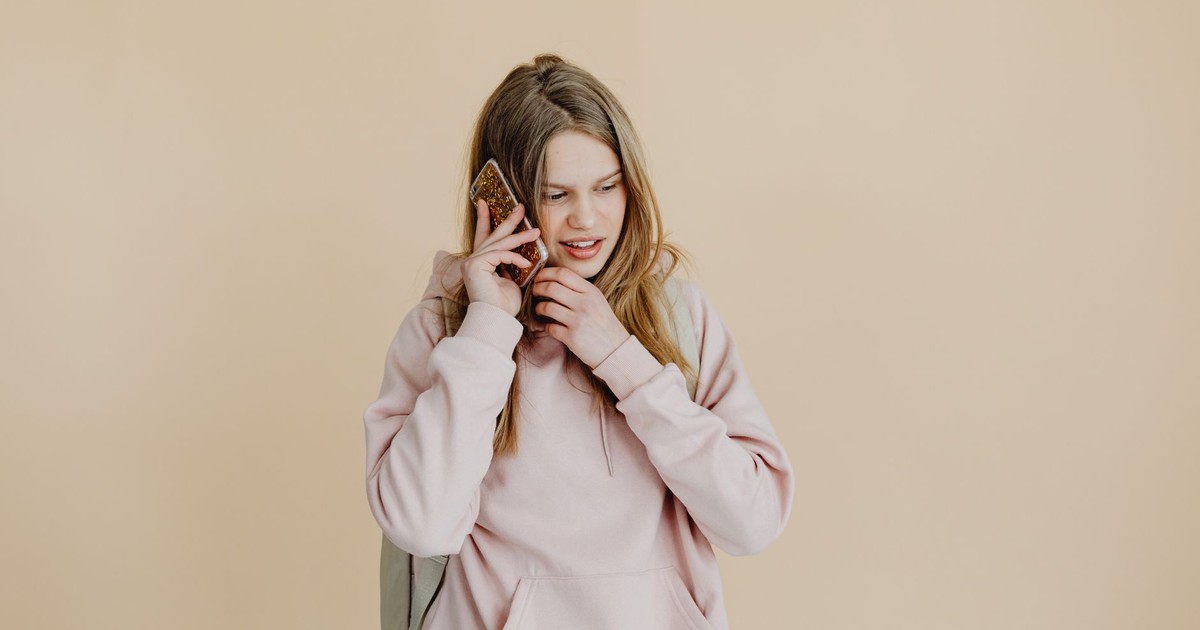 Casual young woman in pink hoodie talking on the phone against a beige background.