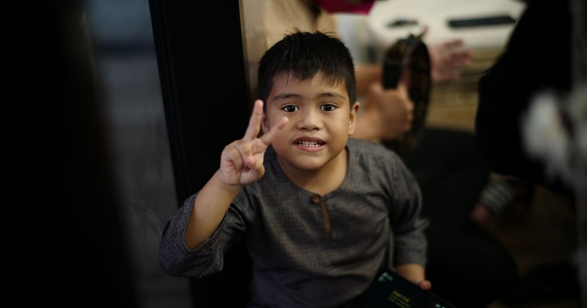 Young boy gesturing peace sign while holding a smartphone indoors, smiling at the camera.