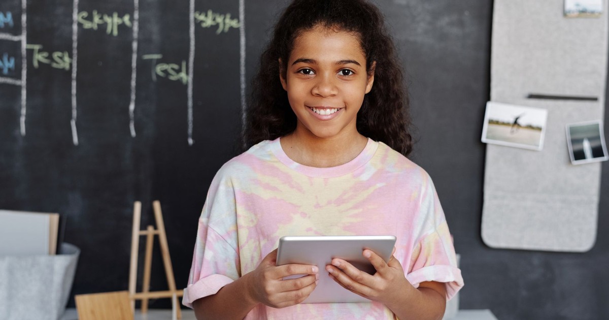 Cheerful girl holding a tablet in a modern classroom with a chalkboard background.