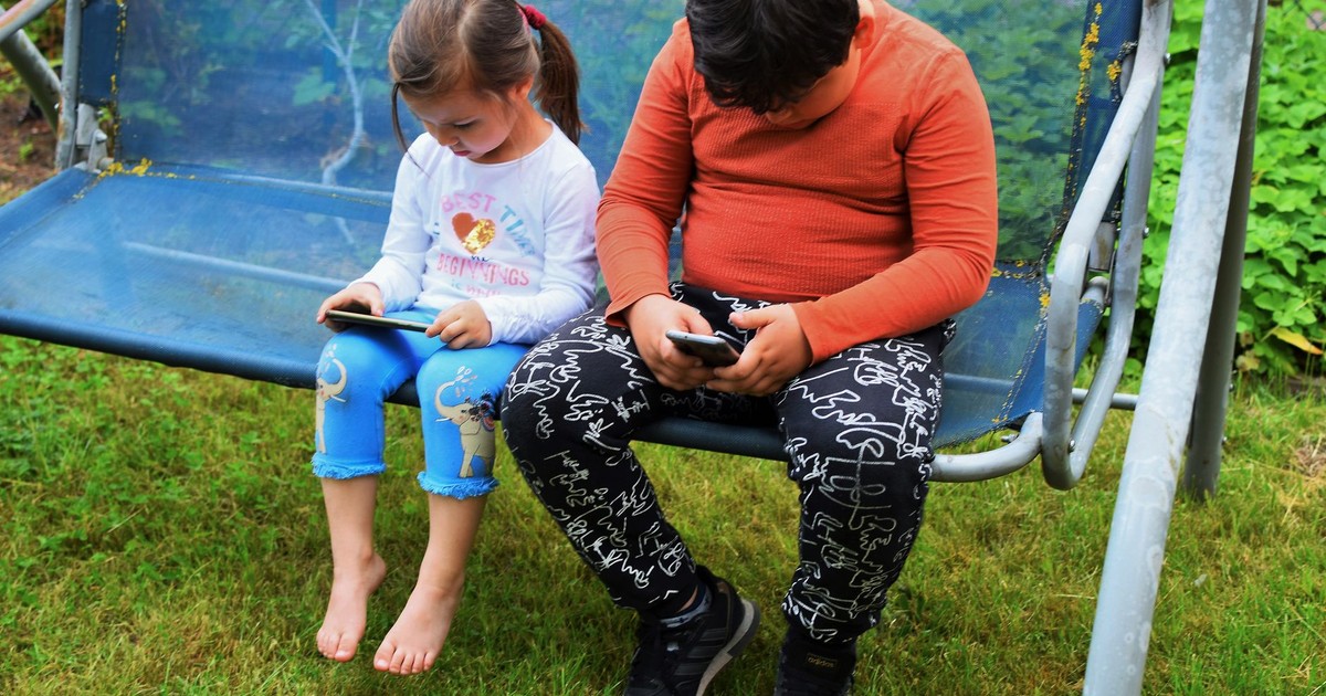 Two children using mobile phones while sitting on a swing outdoors in a garden.