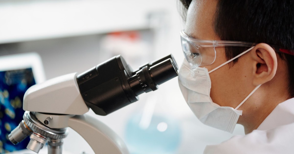 Scientist wearing face mask and goggles examining samples through a microscope in a lab.